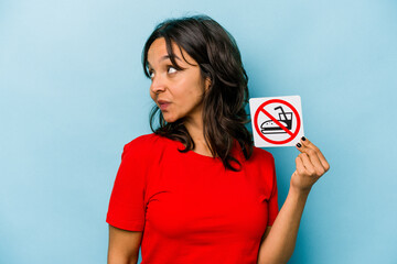 Young hispanic woman holding no eating sign isolated on blue background looks aside smiling, cheerful and pleasant.