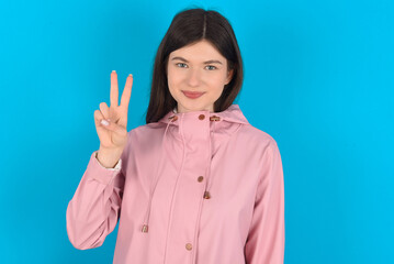 young beautiful Caucasian woman wearing pink raincoat over blue wall showing and pointing up with fingers number two while smiling confident and happy.