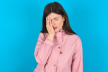 young beautiful Caucasian woman wearing pink raincoat over blue wall Yawning tired covering half face, eye and mouth with hand. Face hurts in pain.