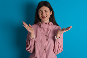 Indignant young beautiful Caucasian woman wearing pink raincoat over blue wall gestures in bewilderment, frowns face with dissatisfaction.