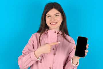 Smiling young beautiful Caucasian woman wearing pink raincoat over blue wall Mock up copy space. Pointing index finger on mobile phone with blank empty screen