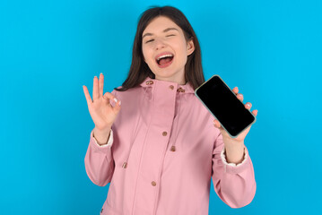 young beautiful Caucasian woman wearing pink raincoat over blue wall holding in hands cell showing ok-sign