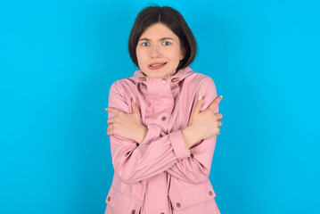 Desperate young beautiful Caucasian woman wearing pink raincoat over blue wall trembles and feels cold, hugs oneself to warm up or feels scared notices something terrifying.