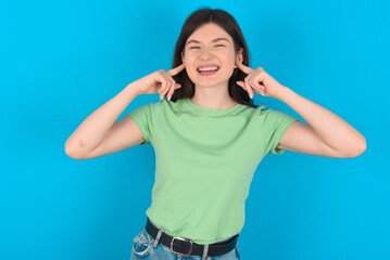 Happy young beautiful Caucasian woman wearing green T-shirt over blue wall ignores loud music and...