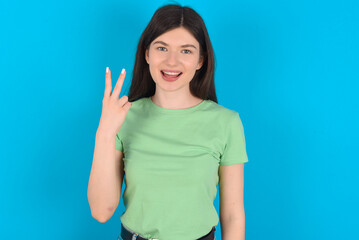 Fototapeta premium young beautiful Caucasian woman wearing green T-shirt over blue wall smiling and looking friendly, showing number two or second with hand forward, counting down