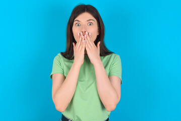 young beautiful Caucasian woman wearing green T-shirt over blue wall keeps hands on mouth, looks with eyes full of disbelief, being puzzled with amount of work