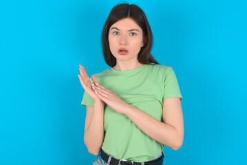 Surprised emotional young beautiful Caucasian woman wearing green T-shirt over blue wall rubs palms and stares at camera with disbelief
