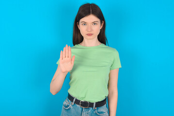 Fototapeta na wymiar young beautiful Caucasian woman wearing green T-shirt over blue wall shows stop sign prohibition symbol keeps palm forward to camera with strict expression