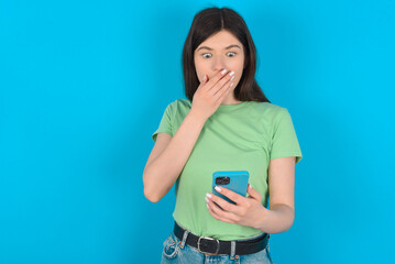 young beautiful Caucasian woman wearing green T-shirt over blue wall being deeply surprised, stares...