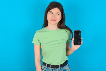 young beautiful Caucasian woman wearing green T-shirt over blue wall holds new mobile phone and looks mysterious aside shows blank display of modern cellular