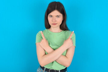 Serious young beautiful Caucasian woman wearing green T-shirt over blue wall crosses hands and points at different sides hesitates between two items. Hard decision concept