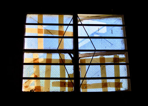 A broken window with hurricane tape reinforcement in a dark room in Havana, Cuba. With an ironic blue sky in the background.
