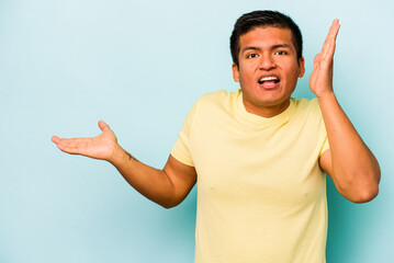 Young hispanic man isolated on blue background holds copy space on a palm, keep hand over cheek. Amazed and delighted.