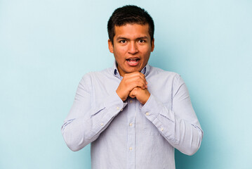Young hispanic man isolated on blue background praying for luck, amazed and opening mouth looking to front.