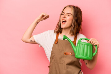 Young gardener woman holding watering can isolated on pink background raising fist after a victory, winner concept. © Asier