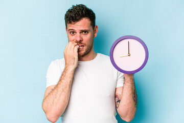 Young caucasian man holding a clock isolated on blue background biting fingernails, nervous and very anxious.