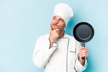 Young caucasian chef man holding flying pan isolated on blue background looking sideways with doubtful and skeptical expression. - obrazy, fototapety, plakaty