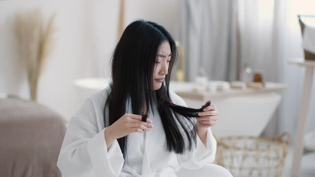 Young upset asian woman looking at her split ends and frowning face, feeling displeased of her hairstyle condition