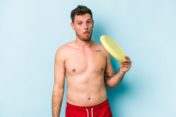 Young caucasian man playing with frisky isolated on blue background shrugs shoulders and open eyes...