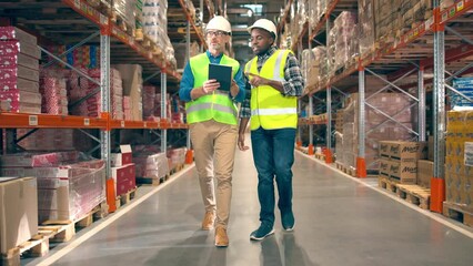 African American male worker walking in retail warehouse in rows with goods in boxes wearing hard hat talking with Caucasian manager checking products stock and inventory typing on tablet Construction