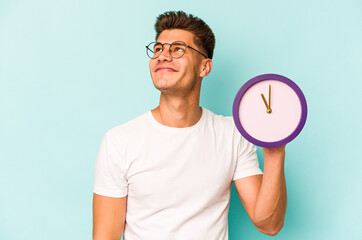 Young caucasian man holding a clock isolated on blue background dreaming of achieving goals and...