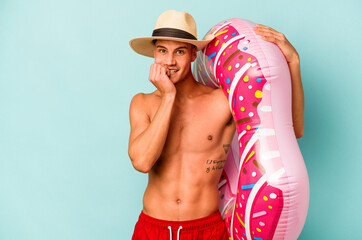 Young caucasian man holding an inflatable donut isolated on blue background biting fingernails, nervous and very anxious.