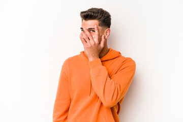 Young caucasian man isolated on white background blink at the camera through fingers, embarrassed covering face.