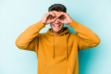 Young caucasian man isolated on blue background showing okay sign over eyes