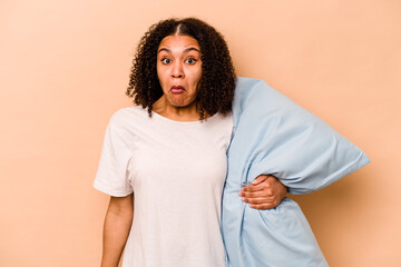 Young African American woman holding a pillow isolated on beige background shrugs shoulders and...