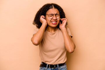 Young African American woman isolated on beige background covering ears with hands.