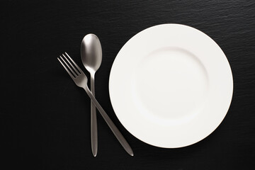 White plate on a black background. Empty white plate, fork, spoon on black slate, flat lay.