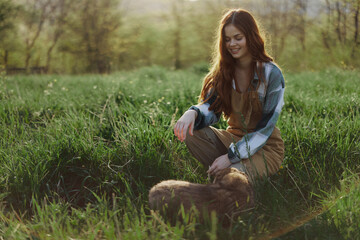 Young redheaded woman playing in nature with her dog in sunny park at sunset in summer - obrazy, fototapety, plakaty