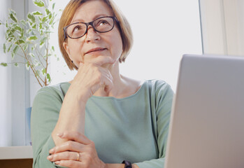 Thoughtful elderly blonde woman in eyeglasses with laptop, looking up, thinking over task, analyzing solutions, options solving problem, dreaming, visualizing, imagining in her head sitting indoor. - Powered by Adobe