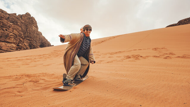 Young woman posing as sand dune surfing wearing bisht - traditional Bedouin coat. Sandsurfing is one of the attractions in Wadi Rum desert