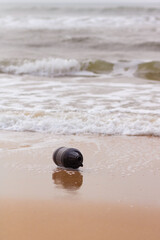 An empty plastic black bottle at the beach being dragged by the ocean waves on the sand. Water and sea pollution concept. Vertical composition