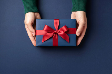 Female's hands holding blue gift box with red bow on blue background. Christmas greeting card.