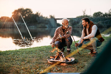 Happy senior father and his son enjoy in preparing fish on bonfire while camping at sunset.