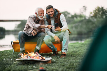 Happy senior father and his son using smart phone by bonfire while camping in nature.