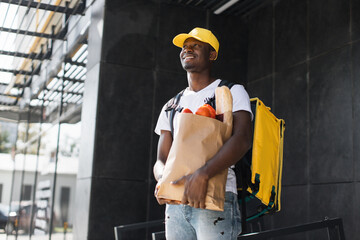 Food delivery African man holding paper bag with organic fresh vegetables with yellow cap and...