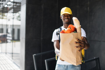 Delivery concept. African American courier standing with a package with fresh food from the store. Smiling African delivery man holding paper bag with organic vegetables.