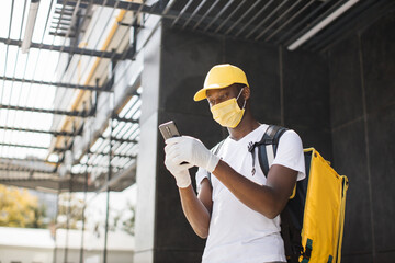 African American male courier food delivery in face mask with yellow thermal backpack looks around street uses smartphone navigate working during covid-19 pandemic.