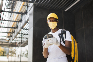African American male courier food delivery in face mask with yellow thermal backpack looks around street uses smartphone navigate working during covid-19 pandemic.