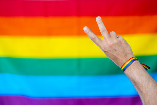Mano haciendo el s&iacute;mbolo de la paz con pulsera de colores del colectivo lgtbi, con la bander lgtbi de fondo