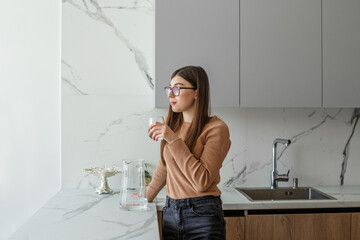 Young adult girl drinks clean water from glass in kitchen. Concept of healthy lifestyle.
