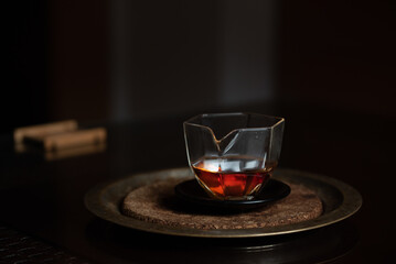 A small Chinese glass jug stands on a dark table against a dark background. There is caramel-colored tea in the pot. The pot is on a tray.