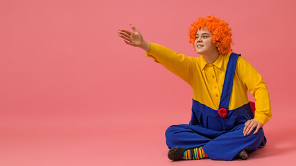 laughing clown in a wig and a yellow-blue suit sits cross-legged and gestures with his hand to the...