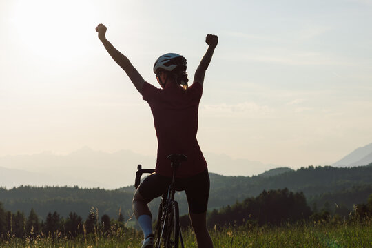 Woman on a road racing bike stopping at a green glade, celebrating with arms raised while looking at an amazing mountain panorama at sunset