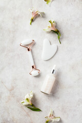 White jade face roller, gua sha, serum with flowers on stone table. Face skin care tools concept.