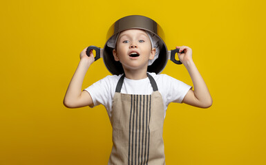 Funny little chef in an apron holding a pot on his head on an isolated yellow background. the concept of helping mom in the kitchen