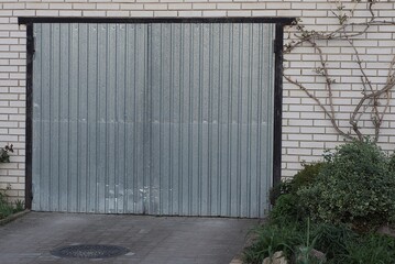 white brick garage with a gray closed metal gate on the street overgrown with vegetation